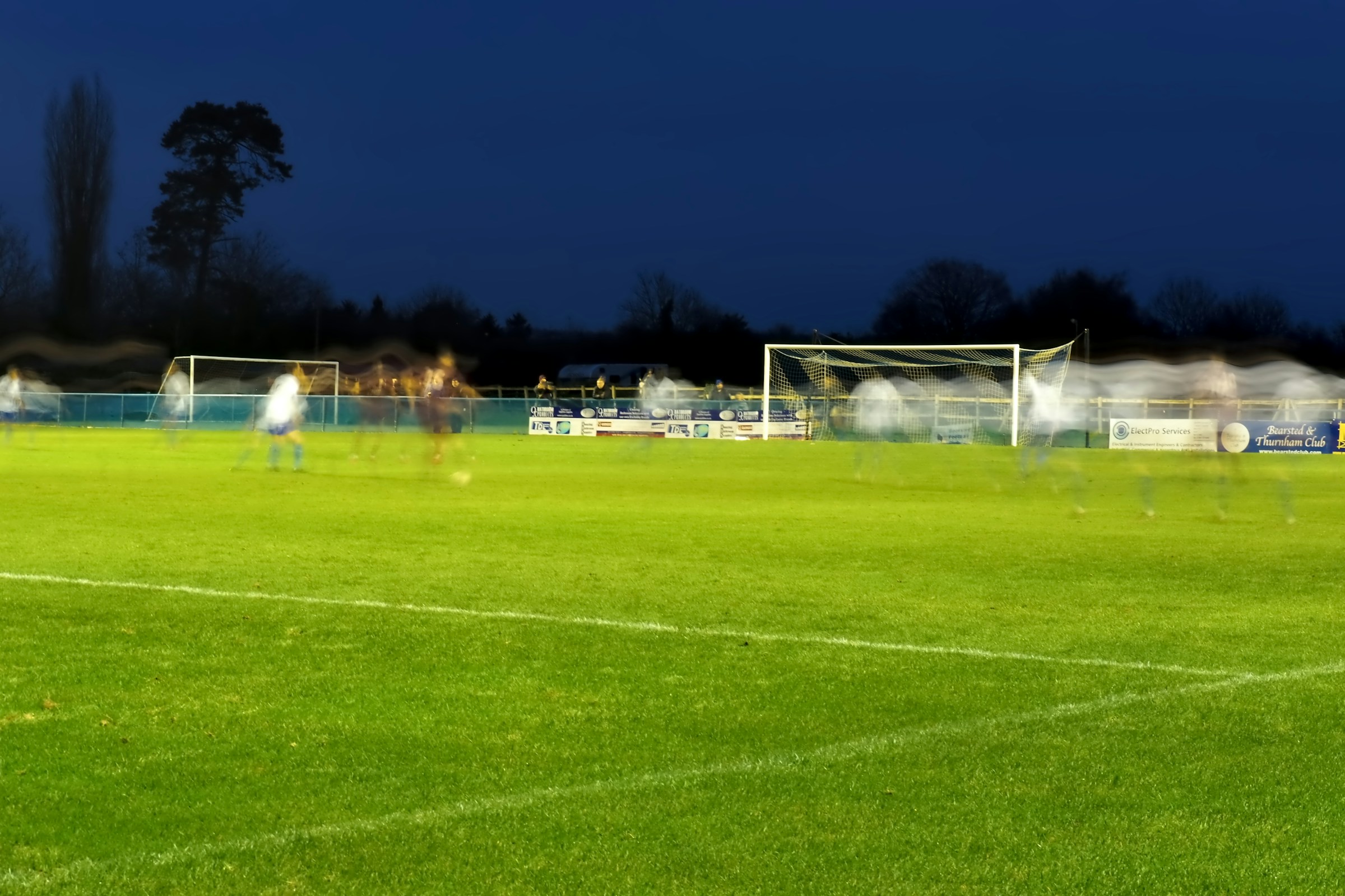 An empty community soccer field lit by floodlights at dusk, goal posts and pitch markings visible under the evening sky.