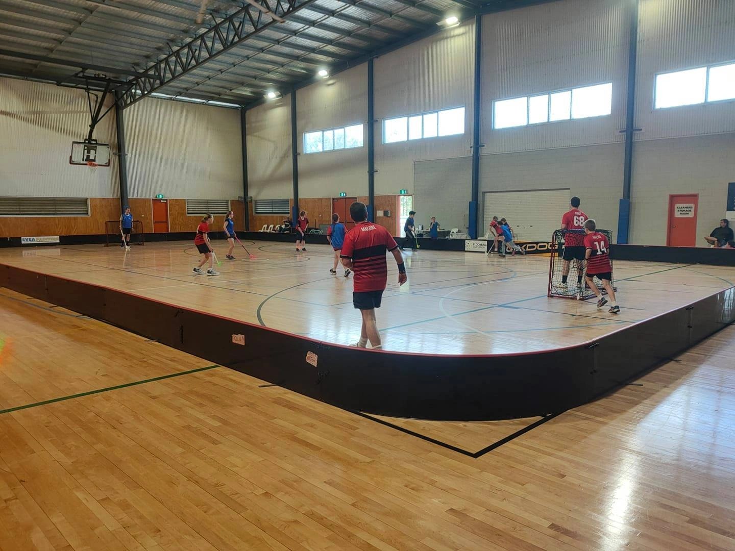 A floorball match underway on the indoor court, players and a goal visible inside the rink boards.