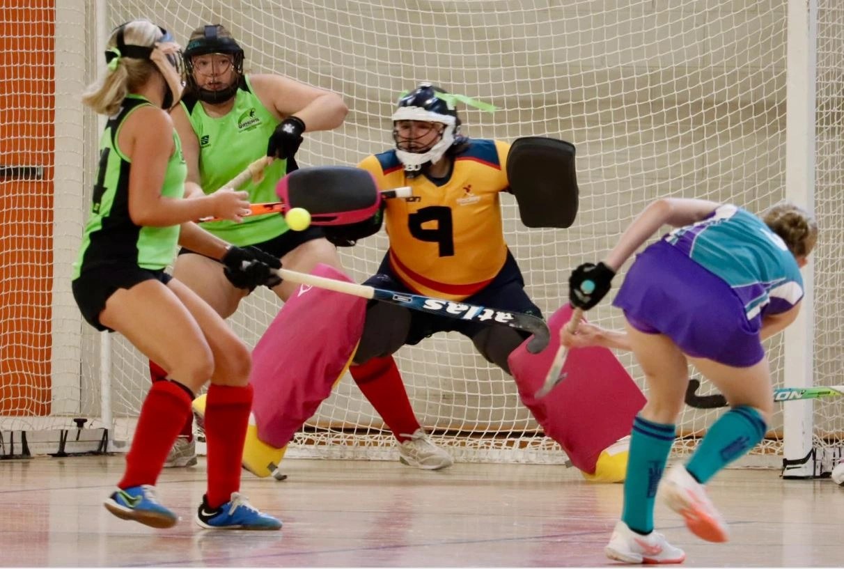 Players in a fast indoor hockey match on the sprung court, goalkeeper defending the net.